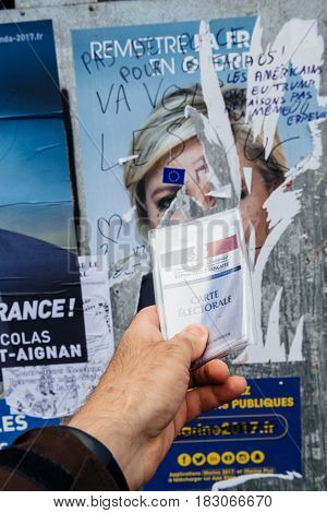 STRASBOURG FRANCE - APR 23 2017: French voter registration card held by male hand in front of official campaign poster of Marine Le Pen candidate for the 2017 French presidential elections posted outside a polling station