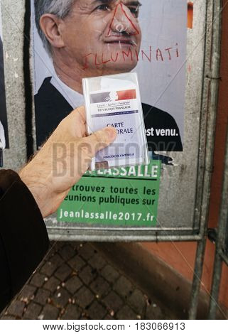 STRASBOURG FRANCE - APR 23 2017: French voter registration card held by male hand in front of official campaign poster of Jean Lassalle candidate for the 2017 French presidential elections posted outside a polling station