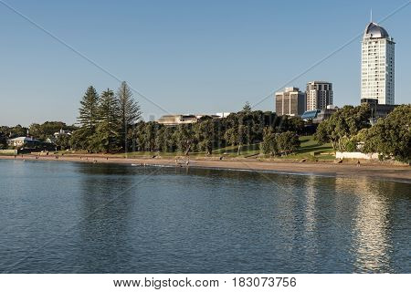 Auckland New Zealand - March 2 2017: South shore of Tapapuna Beach shows park highrise and canoe rowers under blue sky with reflecting sea water.