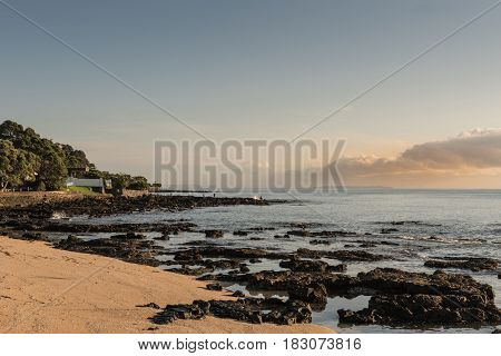 Auckland New Zealand - March 2 2017: Black volcanic north shore of Tapapuna Beach shows upcoming storm clouds over Hauraki Gulf. Some shore vegetation and fishermen.
