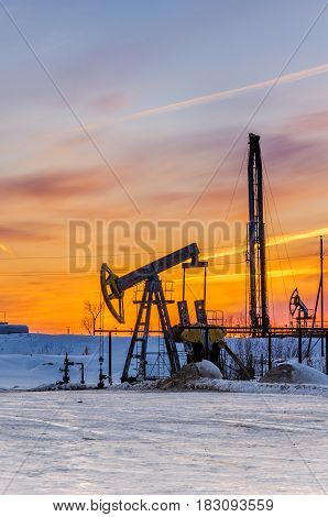 Pump jack, wellhead and oil rig during sunset in the oilfield. Winter period. Oil and gas concept.