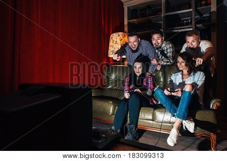 Group of youth have fun playing video game, competition at home. Two girls with joysticks on couch, three guys cheer emotionally, free space