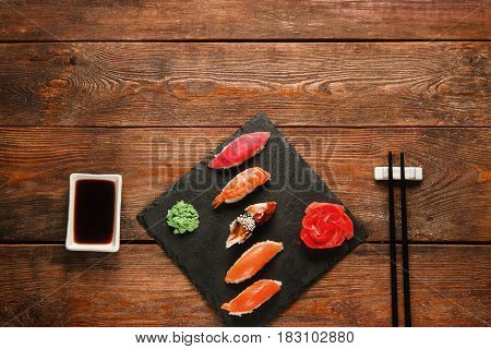 Tasty delicious set of five sushi nigiri served on black slate with ginger and wasabi, on wooden rustic table, flat lay. Japanese cuisine, dark background with free space.