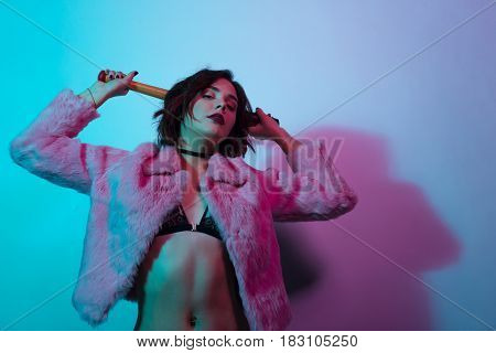 Young female in black bra and pink fur holding batt and looking at camera in neon lights.