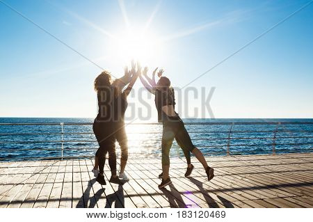 Silhouettes of young beautiful sportive girls giving highfive near sea at sunrise.