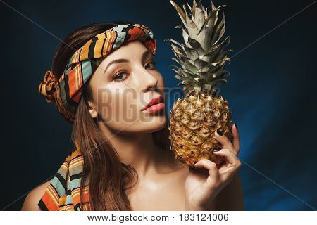 Gorgeous naked woman with colourful bandana on head, holding pineapple, looking straight at camera.