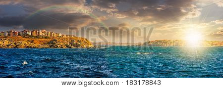 composite summer seascape. panoramic view of old resort town under the rainbow on a rocky cliff above the seashore. blue and calm water in the sea at sunset;