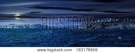 composite summer seascape. panoramic view of old resort town on a rocky cliff above the seashore. blue and calm water in the sea at night in full moon light