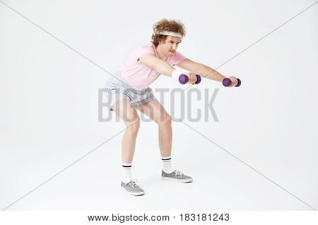 Young skinny man does squats holding light dumbbells, building leg muscles. Looking motivated and concentrated