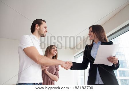 Real estate agent showing apartment to young man and woman, cheerful smiling couple shaking hands with friendly realtor holding papers, client and businesswoman handshaking, business greeting