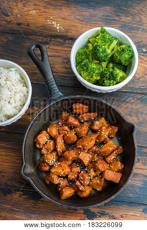 Chicken teriyaki pan-fried in a cast iron skillet with garnish of rice and broccoli in white bowls. Wooden dark rustic table top view vertical photo.