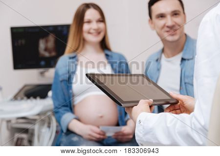 Enjoying friendly conversation with patients. Skillful qualified professional practitioner working in the clinic and using gadget while young couple enjoying appointment