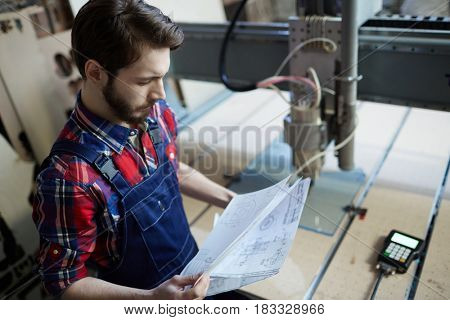 Serious engineer looking at technical sketches by workplace