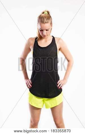 Attractive young fitness woman in black tank top and neon yellow shorts, arms on hips. Studio shot on gray background.