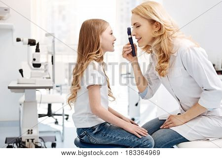 Pretty eyes. Trained competent nice ophthalmologist employing ophthalmoscope for observing girls eyes while giving a consultation