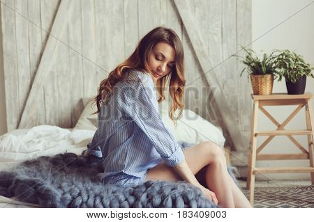 young woman in pajama wake up in the morning in cozy scandinavian bedroom and sitting on bed with houseplants on background. Casual lifestyle in modern interior