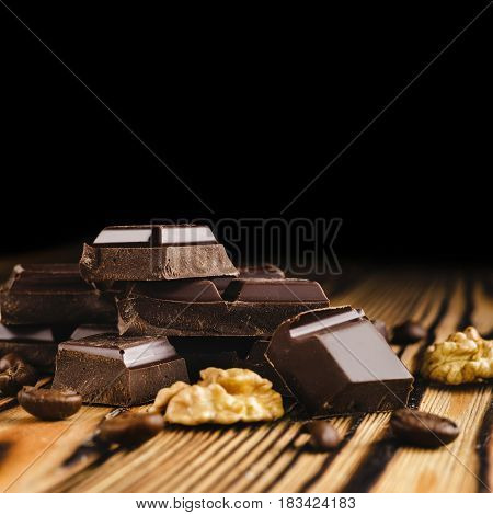 Pieces of bitter dark chocolate cubes on heap walnut core and coffee bean on wooden background close-up view.