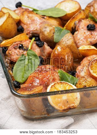 Close up view of chicken thighs with potatoes, lemon and black olives, cooked in oven on gray concrete background. Baked chicken leg quarter in heat-proof glass.