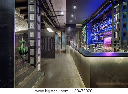 Luminous restaurant in a loft style with brick and concrete walls, column, stairway and a glossy floor. There is a bar rack, shelves with bottles and glasses, fridge, bar equipment, big cactus.