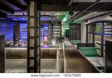 Bar in a loft style with brick and concrete walls, column, glowing lamps and a glossy floor. There is a bar rack, shelves with bottles and glasses, fridge, bar equipment, chairs, tables with sofas.
