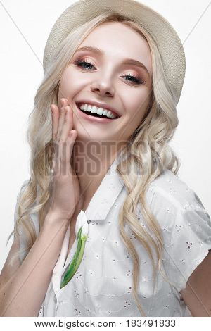 Young smiling girl with curls in a hat. A beautiful model in a white shirt and a brooch-flower. Gentle makeup. Summer light image. Beauty of the face.