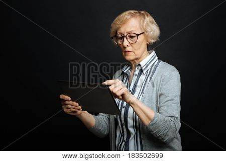 Becoming familiar with modern technologies. Concentrated skilled senior woman demonstrating interest and using gadget while standing isolated in black background
