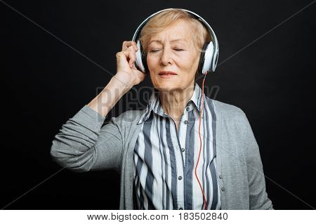Deep in the world of music . Delightful cheerful happy pensioner expressing delight and using headphones while listening to the music and standing against black background