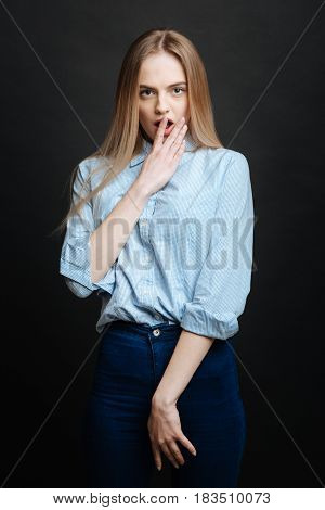 Under impression. Young surprised emotional teenager expressing astonishment and raising hand to the mouth while standing isolated in black background