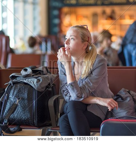 Young blond caucsian woman waiting on airport terminal full of passenegers for her flight to depart.