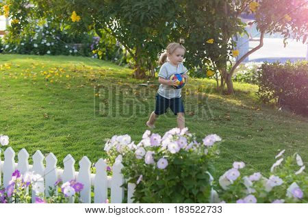 sport game and running. cute baby boy happy small little child with long blond hair holds colorful ball on green grass in garden with blossoming flowers on sunny summer day on natural background