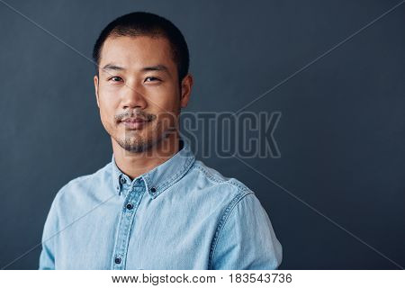 Portrait of a casually dressed young Asian designer smiling confidently while standing against a gray wall in an office