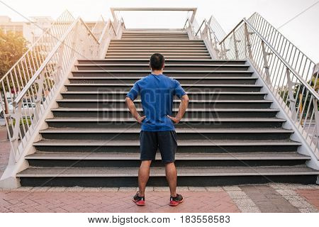 Rearview of a man standing with his hands on his hips at the bottom of a set of stairs while out for a city run