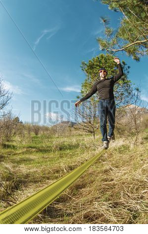 A man, aged with a beard and wearing sunglasses, balances on a slackline in the open air between two trees at sunset on background blue sky