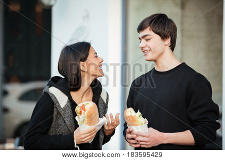 Young brunette woman holding burger and smiling to her brother in the street