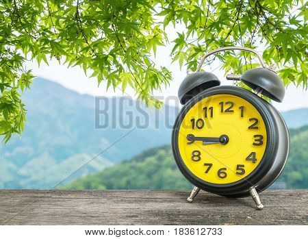Closeup black and yellow alarm clock for decorate show a quarter to nine o c'clock or 8:45 a.m. on blurred leaves and mountain view background