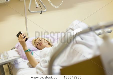 Bedridden female patient lying in hospital bed, using mobile phone while recovering after surgery.