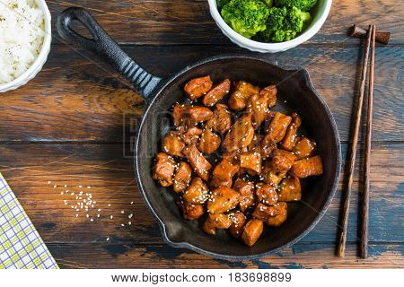 Chicken teriyaki pan-fried in a cast iron skillet with garnish of rice and broccoli in white bowls. Chopsticks wooden rustic table top view.