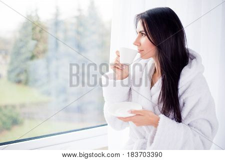Portrait Of A Brunette Young Girl Drinking Coffee At A Hotel Room On Vacation. She Is Dreamy And Rel