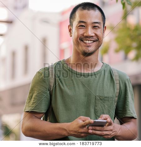 Square portrait of a casually dressed young Asian man smiling while standing outside on a city street sending a text message on his cellphone