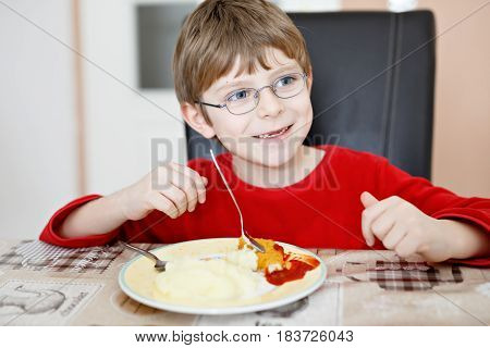 Adorable little school boy eating potato mash and chicken breast indoor. Blond child in domestic kitchen or in school canteen Cute kid and healthy food
