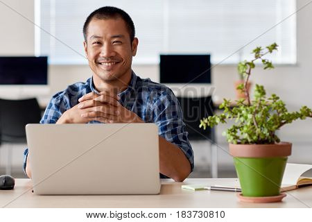 Young Asian entrepreneur smiling confidently while sitting alone at a desk in a modern office working on a laptop, with greenery around him