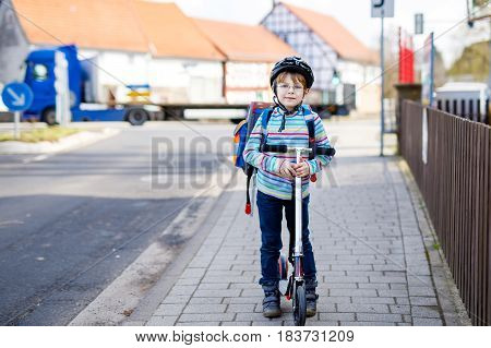 Active school kid boy in safety helmet riding with his scooter in the city with backpack on sunny day. Happy child in colorful clothes biking on way to school. Safe way for kids outdoors to school.