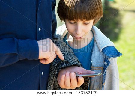 Father pointing at smartphone and boy looking with surprise and interest. Father with son using smart phone outdoor together. Young boy with his father in spring park.