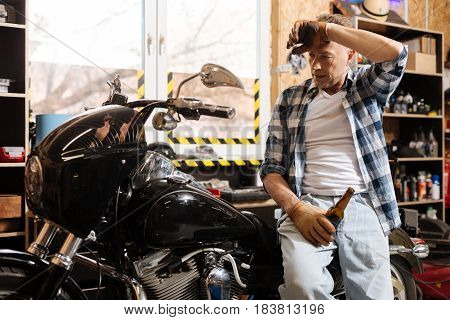 Time to appreciate things. Diligent relaxed amateur serviceman taking some rest and drinking a bear after bringing the bike in perfect condition
