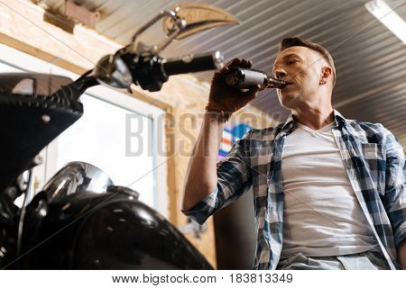 A little cheer up. Reliable delighted mature guy having a little celebration on finishing the reparation of a great bike and drinking some beer