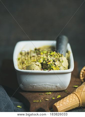 Homemade pistachio ice cream in ceramic mold with metal scooper, crashed pistachio nuts and waffle cones over concrete background, copy space, vertical composition, selective focus