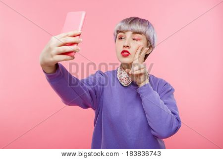 Close-up portrait of beautiful dollish girl with short light violet hair wearing lilac sweater making selfie over pink background. Copy space.