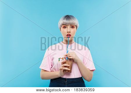 Close up portrait of beautiful dollish girl with short light violet hair in pink tshirt drinking juice over blue background. Copy space.