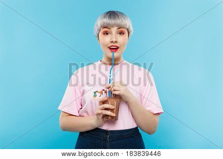 Close up portrait of cheerful smiling beautiful dollish girl with short light violet hair in pink tshirt drinking juice over blue background. Copy space.