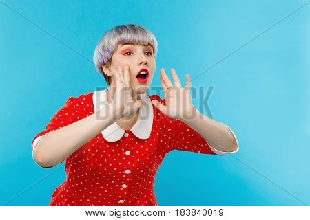 Close up portrait of beautiful dollish girl with short light violet hair wearing red dress shouting over blue background. Copy space.
